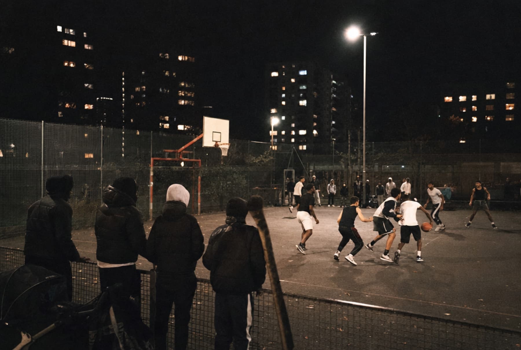 Spectateurs de dos au grillage d’un city stade éclairé, match de basket en cours